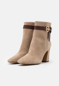 Un par de botines de ante beige con puntera afilada, tacones cuadrados y correas de cuero marrón oscuro sujetas por hebillas doradas.