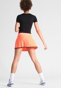 Gonnella da tennis plissettata in gradiente arancione, con una cintura larga. Abbinata a un top nero aderente e calze e scarpe sportive bianche.