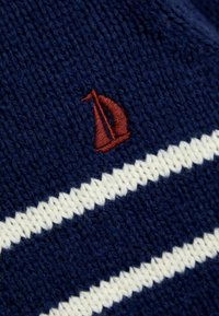 Marinefarbener Strickstoff mit weißen horizontalen Streifen und einem kleinen roten gestickten Segelboot-Logo in der oberen linken Ecke.