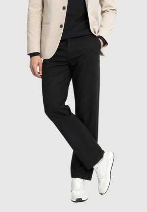 Persona che indossa pantaloni neri, sneakers bianche, camicia nera e blazer beige con una mano in tasca e gambe incrociate.