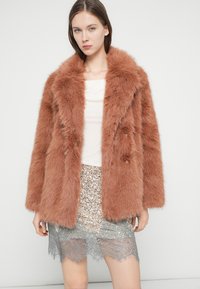 Παλτό από συνθετική γούνα σε απαλό σκουριασμένο χρώμα, με φαρδύ γιακά και διπλό κούμπωμα. Φοράται πάνω από φούστα με παγιέτες και δαντελένια ρίγα.