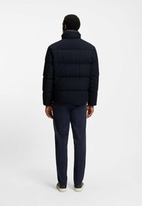 Giacca imbottita navy con tessuto testurizzato, colletto alto e cuciture visibili. Indossata con pantaloni scuri e calzature di colore chiaro.