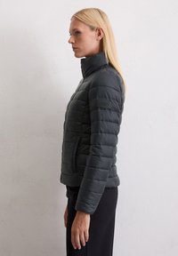 Kvinne med rett, blondt hår som har på seg en svart kviltet jakke med høy krage, stående i profil mot en enkel hvit vegg.