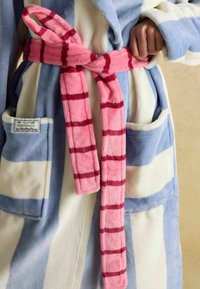 Inimene sidub roosa ruudulisest vööst sinise ja valge triibulise plush-käterätiku ümber, millel on ees taskud ja silt "Joules Hibernate".