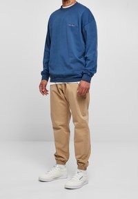 Muž v modrém sweatshirtu s dlouhým rukávem, béžových kalhotách s manžetami a bílých teniskách, stojí před jednobarevným světle šedým pozadím.