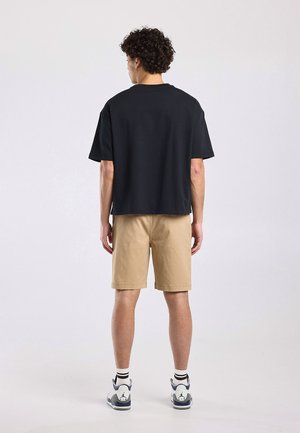 Man met krullend haar staat met zijn rug naar ons toe, draagt een zwart T-shirt met korte mouwen, beige korte broek, witte sokken en witte sneakers.