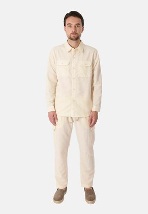 Uomo con capelli corti e scuri e barba, che indossa una camicia crema con bottoni e pantaloni con scarpe slip-on marroni, in piedi davanti a uno sfondo semplice.