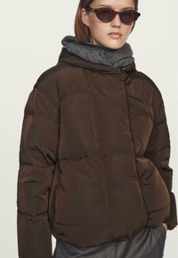 Brown, puffy jacket with a hood, made of textured material. Features a zip closure and side pockets, layered over a dark gray scarf.