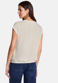 Top beige a maniche corte con scollatura rotonda e orlo elasticizzato, abbinato a jeans blu scuro. Il tessuto appare liscio con minima lucentezza.
