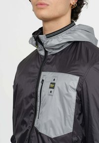 Giacca leggera nera e grigia con chiusura zip, cappuccio regolabile e tessuto testurizzato. Presenta una patch con logo sulla tasca del petto.