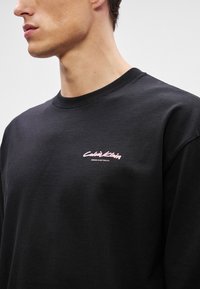 Svart langermet t-skjorte, laget av bomull, med en rosa Calvin Klein-logo på brystet og rund hals. Glatt tekstur.