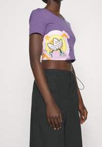 Fioletowy bawełniany crop top z zabawną grafiką postaci na pastelowym tle z chmurami; krótki rękaw, dopasowany krój.