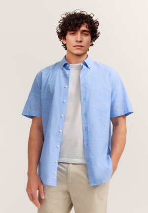 Junger Mann mit lockigem Haar, der ein hellblaues kurzärmeliges Hemd über einem weißen T-Shirt und beige Hose trägt, steht vor einem einfarbigen Hintergrund.