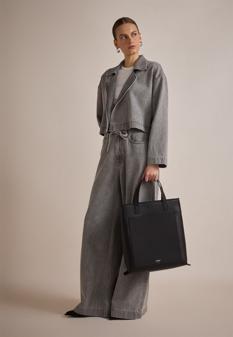 Sac fourre-tout noir avec une texture lisse, de forme rectangulaire et des poignées courtes. Porté avec une veste grise cropped et un pantalon large.