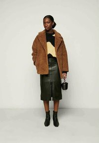 Γυναίκα φορώντας ένα καφέ αρκουδοπαλτό, μαύρο και μπεζ πουλόβερ, σκούρα πράσινη δερμάτινη μίντι φούστα, μαύρες μπότες έως τον αστράγαλο, κρατώντας μια μικρή μαύρη τσάντα.