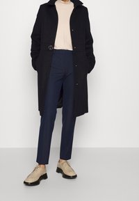 Persona vestida con un abrigo negro sobre una blusa beige, pantalones a medida azul marino y zapatos de cuero beige con cordones, de pie contra un fondo blanco liso.