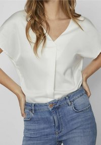 Kvinna som bär en vit V-ringad kortärmad blus instoppad i högmidjade blå jeans, med händerna vilande på höfterna, som visar detaljer av den avslappnade outfiten.