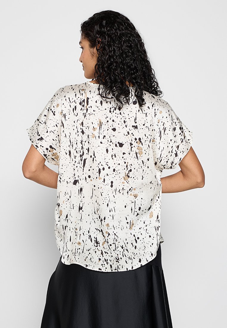 Losse, crèmekleurige blouse met een zwart en goud spettern patroon. Korte mouwen, zachte stof en een afgeronde zoom aan de achterkant.