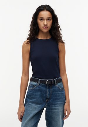 Γυναίκα με μακριά σγουρά μαλλιά, φορώντας μπλε αμάνικη μπλούζα, μπλε τζην και μαύρη ζώνη, στέκεται μπροστά σε απλό λευκό φόντο.