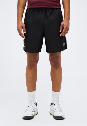Mand iført sorte EA7 mærkede sportsshorts, hvide sokker og hvide sportssko, står op ad en ensfarvet lys baggrund.