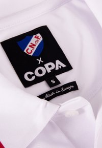 Witte polo met kraag, voorzien van een blauw-rood schildlogo, een zwart label met "COPA" en "Made in Europe," en een kleine witte knop.