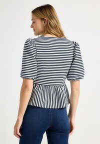 Kvinde iført navyblå og hvide vandrette stribede bluse med pufærmer og rynket talje, kombineret med mørkeblå jeans, set fra siden mod hvid baggrund.