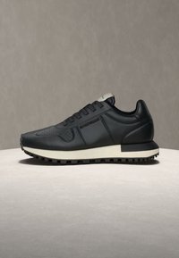 Sneaker noir à tige basse avec une semelle blanche et une semelle extérieure à crampons, featuring des lacets ton sur ton et un marquage embossé subtil sur le côté.