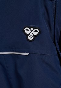 Marinfärgad tyg med en framträdande patch med svart och vitt bi-logo. Inkluderar grå accenter nära sömmen för designdetalj.