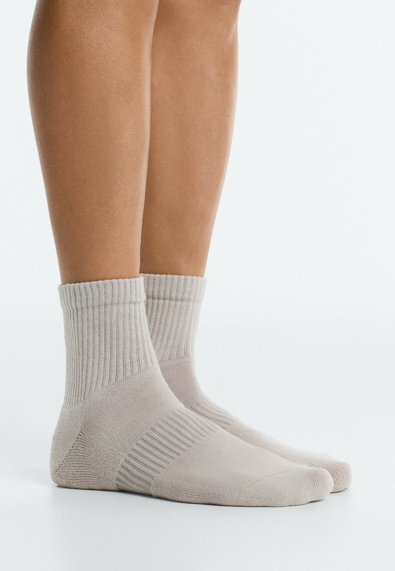 Paire de chaussettes cheville côtelées beige clair portées sur des pieds à peau claire, vues de côté sur fond blanc.