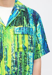 Koszula z krótkim rękawem w żywym zielono-niebieskim abstrakcyjnym wzorze. Lekki materiał z kieszenią na piersi i rozpinanym przodem na guziki.