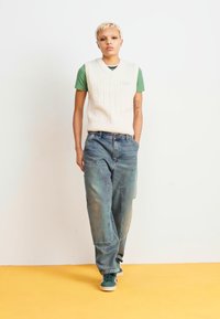 Gilet lavorato a maglia bianco sopra una maglietta verde, abbinato a jeans larghi e sbiaditi blu e sneakers verdi, in piedi davanti a uno sfondo bianco.
