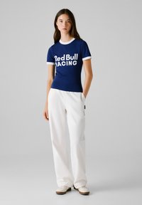 Giovane donna in piedi con una maglietta blu navy "Red Bull Racing", pantaloni bianchi e scarpe chiare su uno sfondo semplice.