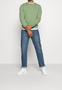 Grön bomulls-tröja med rund halsringning över en vit T-shirt, kombinerad med blå jeans och vita höga sneakers.