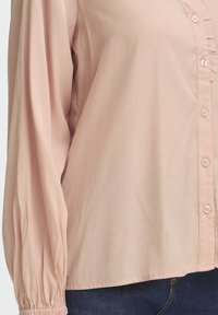 Világos rózsaszín, hosszú ujjú blúz gombos rögzítéssel és fodros díszítéssel elöl. Rugalmas mandzsetták és sima, könnyű anyag.