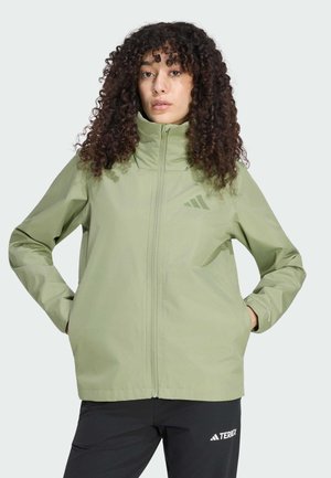 Vrouw draagt een lichtgroene jas met rits, handen in de zakken en zwarte broek met wit Terrex-logo, staand tegen een effen achtergrond.