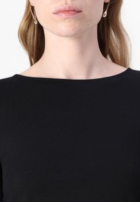 Sorte langermede topp med vid hals, laget av et glatt stoff. Gule sikkerhetsnåløreringer henger fra ørene, og tilfører detaljer.