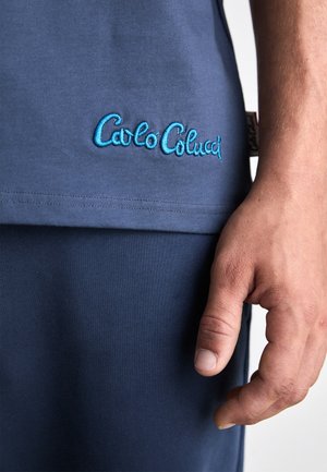 Zbliżenie na rękę osoby obok brzegu niebieskiego topu z niebieskim haftowanym logo "Carlo Colucci" oraz pasujące niebieskie spodnie.