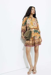 Robe à imprimé floral orange et vert avec des manches courtes, ourlet volanté et une touche de brun. Sac en daim vert olive avec des clous. Sandales à talons beiges avec des lanières.