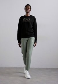 Svart sweatshirt med guldlogotyp, tillsammans med gröna sweatpants med en diskret logotyp, och vita sneakers. Slät tyg, avslappnad passform.