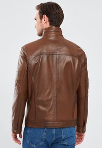 Giacca in pelle marrone con colletto alto, design aderente e dettagli di cucitura sottili. Maniche lunghe con polsini abbottonati e una texture liscia.