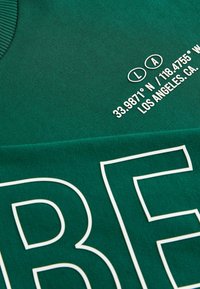 Groene stof met geribbelde halslijn, voorzien van witte tekst en coördinaten die op het oppervlak zijn gedrukt. De textuur is zacht en glad.