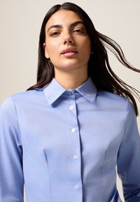 Camicia azzurra chiara con colletto classico, maniche lunghe e una sottile texture del tessuto. Presenta bottoni bianchi lungo il davanti.