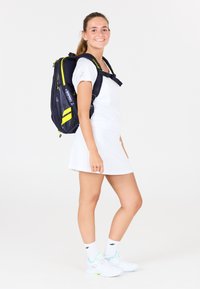 Marineblå og gul rygsæk med flere rum og mesh-detaljer, bæret af en person i et hvidt atletisk outfit og sko.