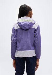 Άτομο με κοτσίδες που φοράει αδιάβροχο μπουφάν Columbia σε μοβ και ανοιχτό γκρι χρώμα και σκούρο παντελόνι, κοιτάζοντας προς τα πίσω με φόντο απλό.