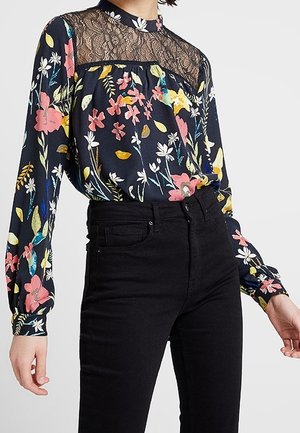 Kobieta w czarnej, kwiecistej bluzce z koronkowym karczkiem i wysokim kołnierzem, w połączeniu z wysokim stanem czarnych dżinsów.