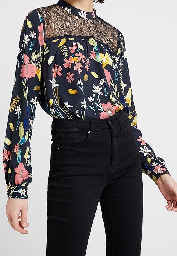 Femme portant un chemisier noir à fleurs avec un empiècement en dentelle et un col haut, associé à un jean noir taille haute.