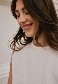 Haut tricoté blanc sans manches avec un col côtelé. Texture douce, lignes épurées, sans motifs visibles. Le modèle a les cheveux bruns et les yeux fermés.