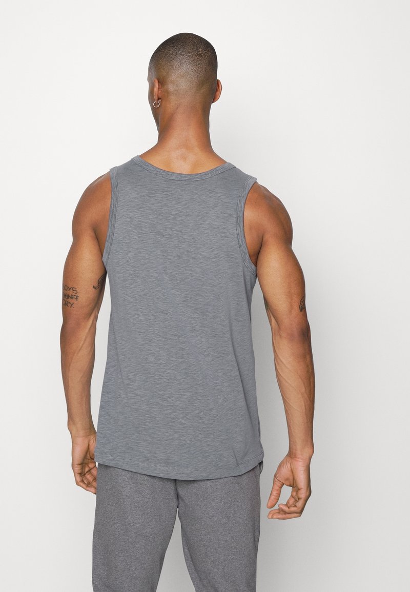Homme vu de dos portant un débardeur gris et un pantalon gris, debout devant un fond clair uni.