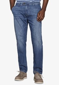 Hombre con vaqueros de corte recto azul y las manos en los bolsillos, calzando zapatos taupe con cordones, sobre un fondo blanco.