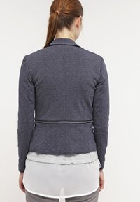 Marinblå skräddarsydd blazer med en figurnära design, dragkedjedetalj vid midjan och en strukturerad yta. Bärs över en ljusgrå skjorta med en genomskinlig nederkant.